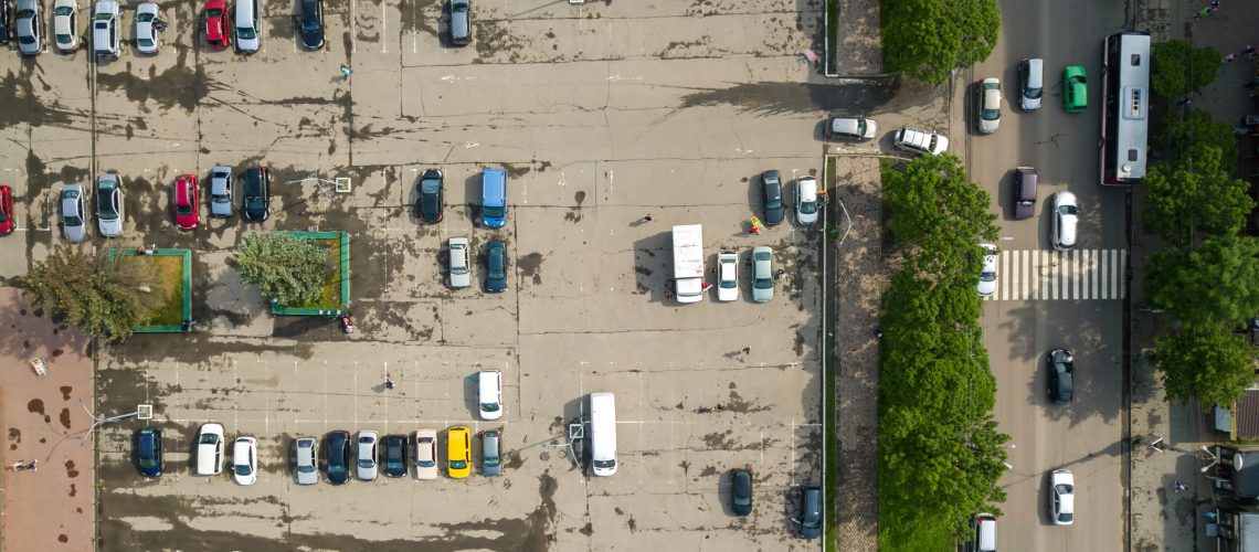 Top down aerial view of busy street with moving cars traffic and big parking lot with many parked vehicles.